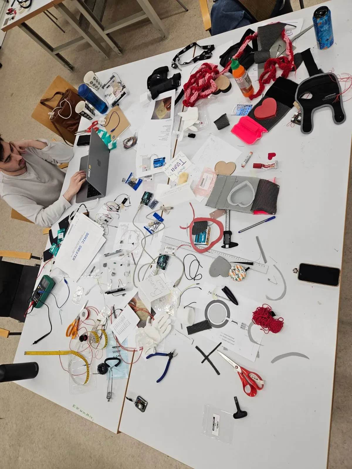 Messy table full of electronic components and fabric pieces, with Bruno Norrå sitting before a laptop.
